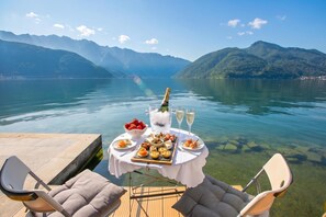 Ein Tisch reichhaltig gedeckt mit Speisen und Getränken, mit Blick auf einen See und dahinter Berge.