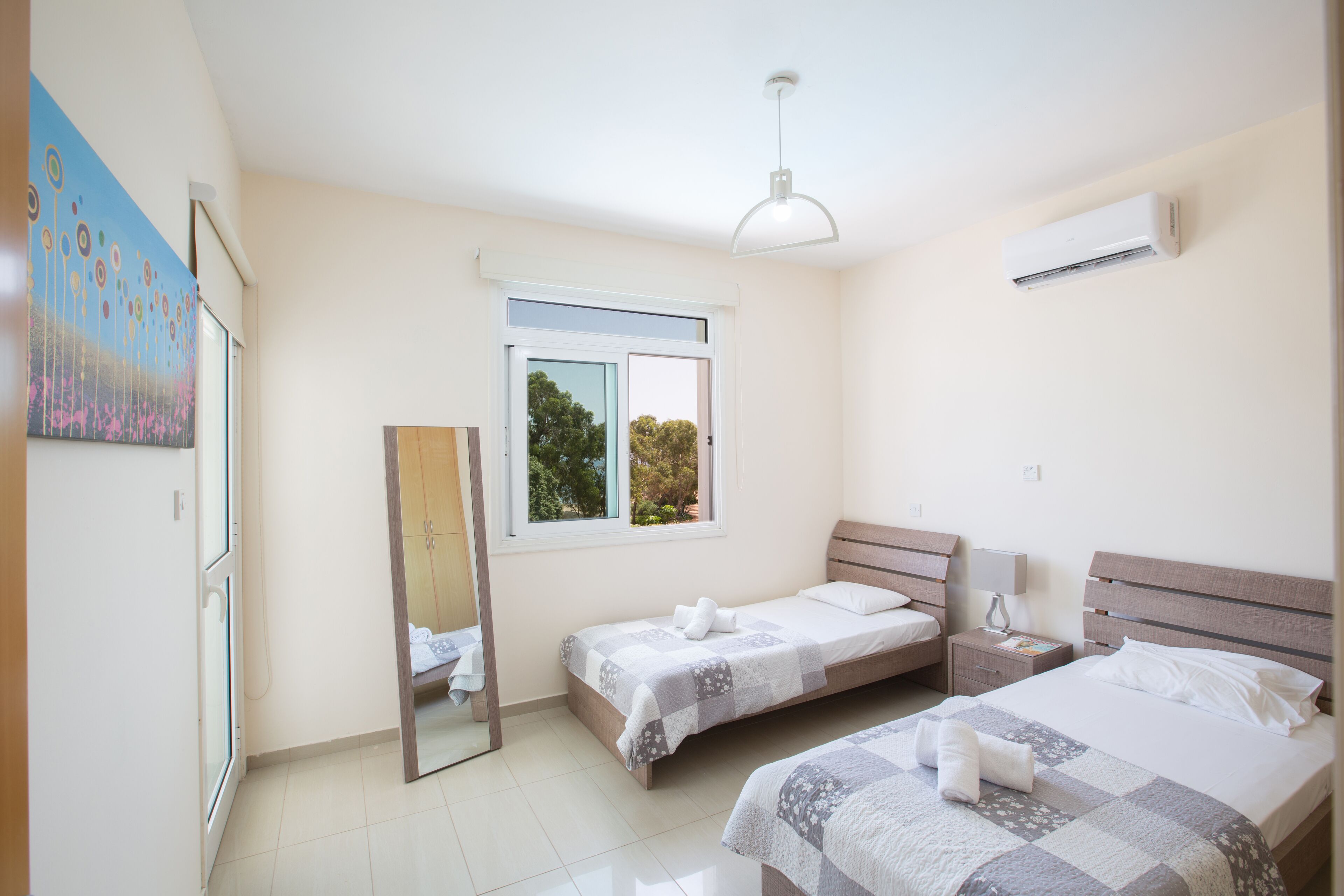 A bedroom with two single beds, a window, a mirror, and an air conditioning unit.