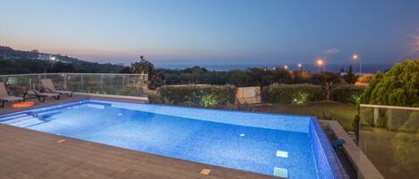 A swimming pool with a wooden deck, surrounded by a glass railing and a view of the landscape.
