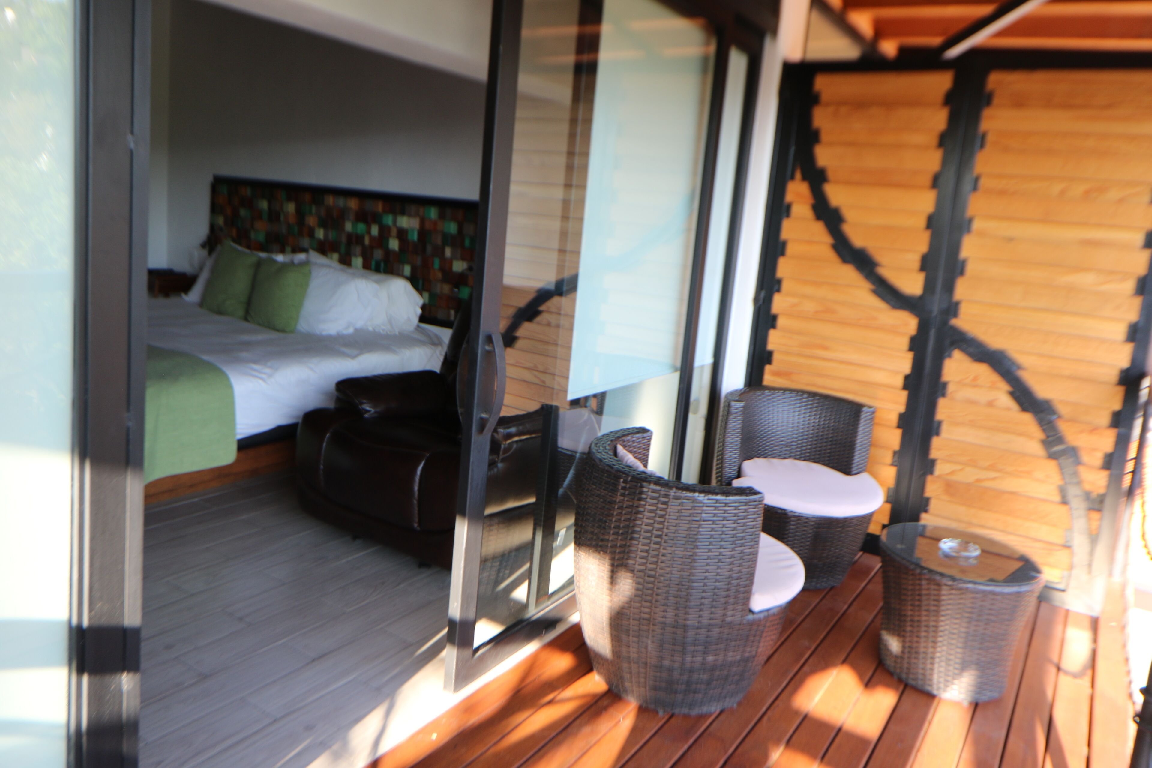 A bedroom with a bed, green pillows, and a suitcase. There is a wicker chair and a small table on a wooden deck.