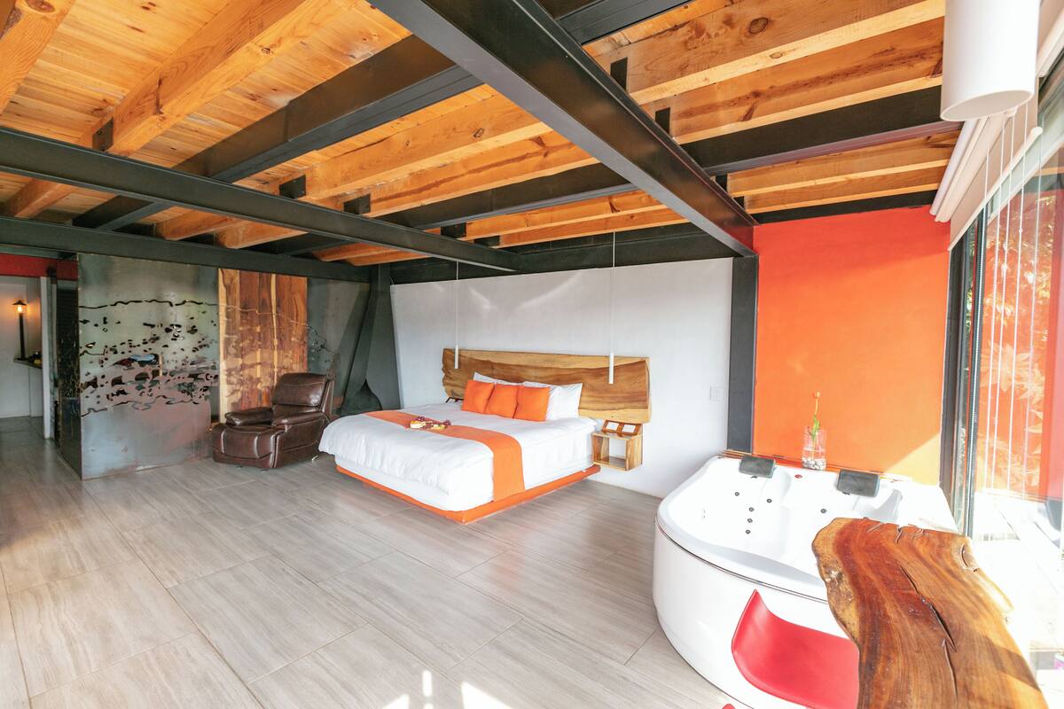 Un dormitorio moderno con techo de madera, una cama grande con ropa de cama naranja, una silla roja, una mesita de noche de madera y una bañera blanca.
