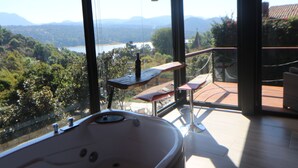 Un baño moderno con una bañera grande, una mesa de madera y una copa de vino en un balcón con vistas a un paisaje pintoresco.