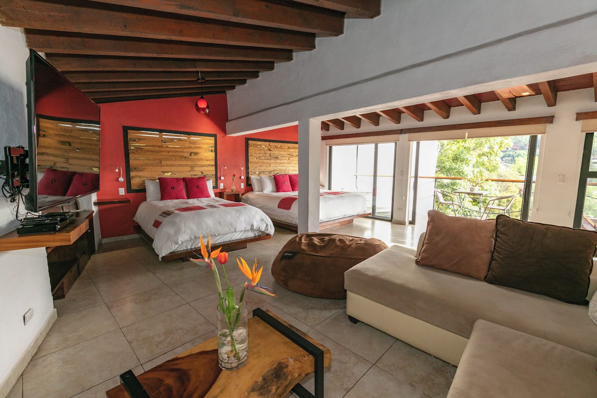 Una habitación de hotel moderna con una cama grande, un sofá, una mesa de centro de madera y vistas al exterior.