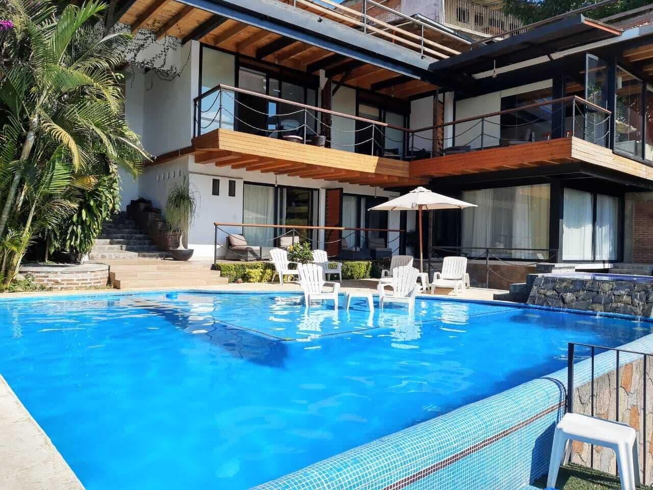 Un área de piscina moderna con una piscina azul cristalina, sillones blancos y una terraza de madera.