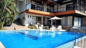 Un área de piscina moderna con una piscina azul cristalina, sillones blancos y una terraza de madera.