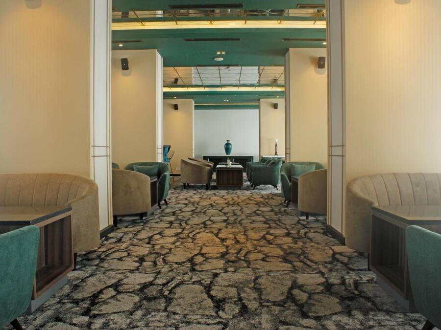 Un vestíbulo de hotel con alfombra estampada, sillones verdes, una mesa central y un jarrón en la pared de fondo.