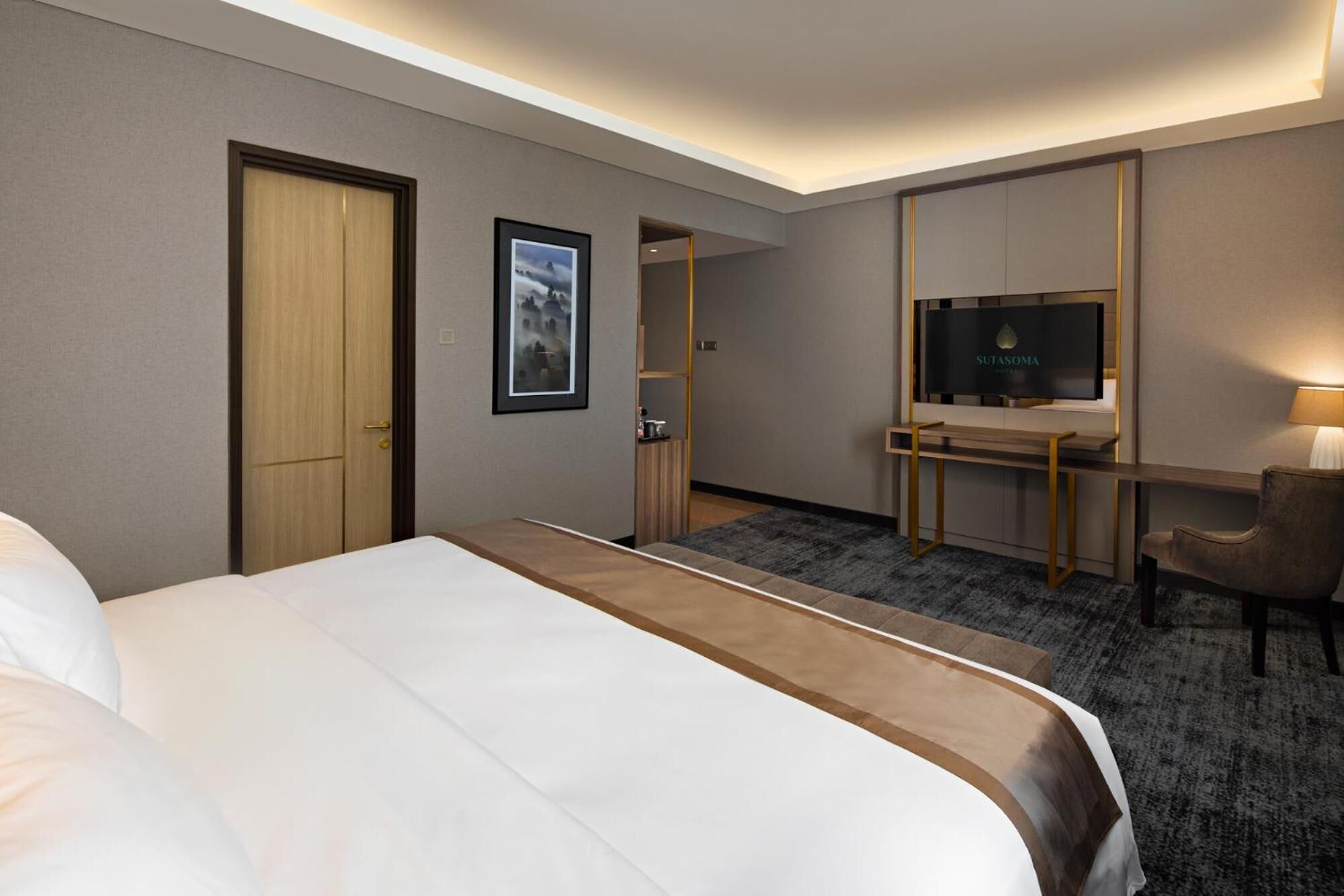 A hotel room with a large bed, a desk with a flat-screen TV, a chair, a door, and a framed picture on the wall.