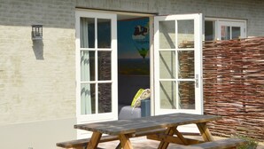 A wooden picnic table outside a building with a glass door, a wicker fence, and a banner with a penguin illustration. - Holiday Home in Haaren Near De Efteling (Haaren)