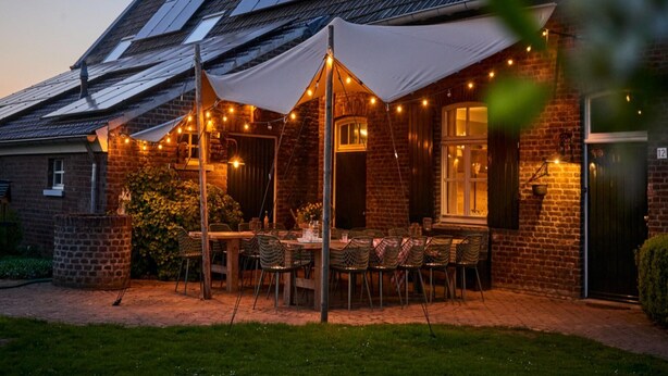 A brick building with a sloped roof, a covered outdoor seating area with tables and chairs, and string lights. - Sprawling Farmhouse in Baarlo With Hot Tub, Private Garden (Baarlo)
