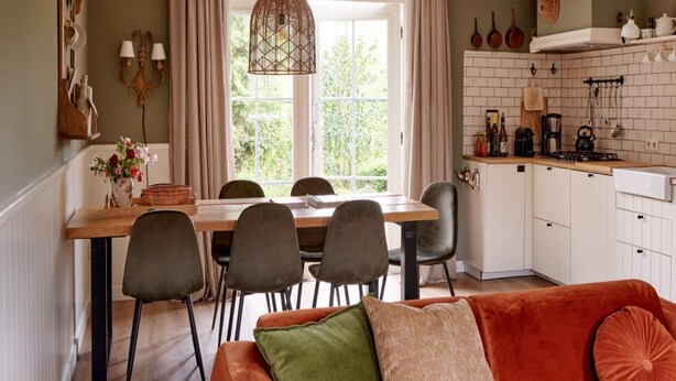 Una cucina accogliente con tavolo da pranzo, sedie e un divano. Si vede una finestra con tende, un lampadario e pensili da cucina.