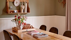 Zona pranzo con tavolo in legno, sedie, un vaso con fiori, un cesto di vimini e una mensola con piatti e bottiglie.