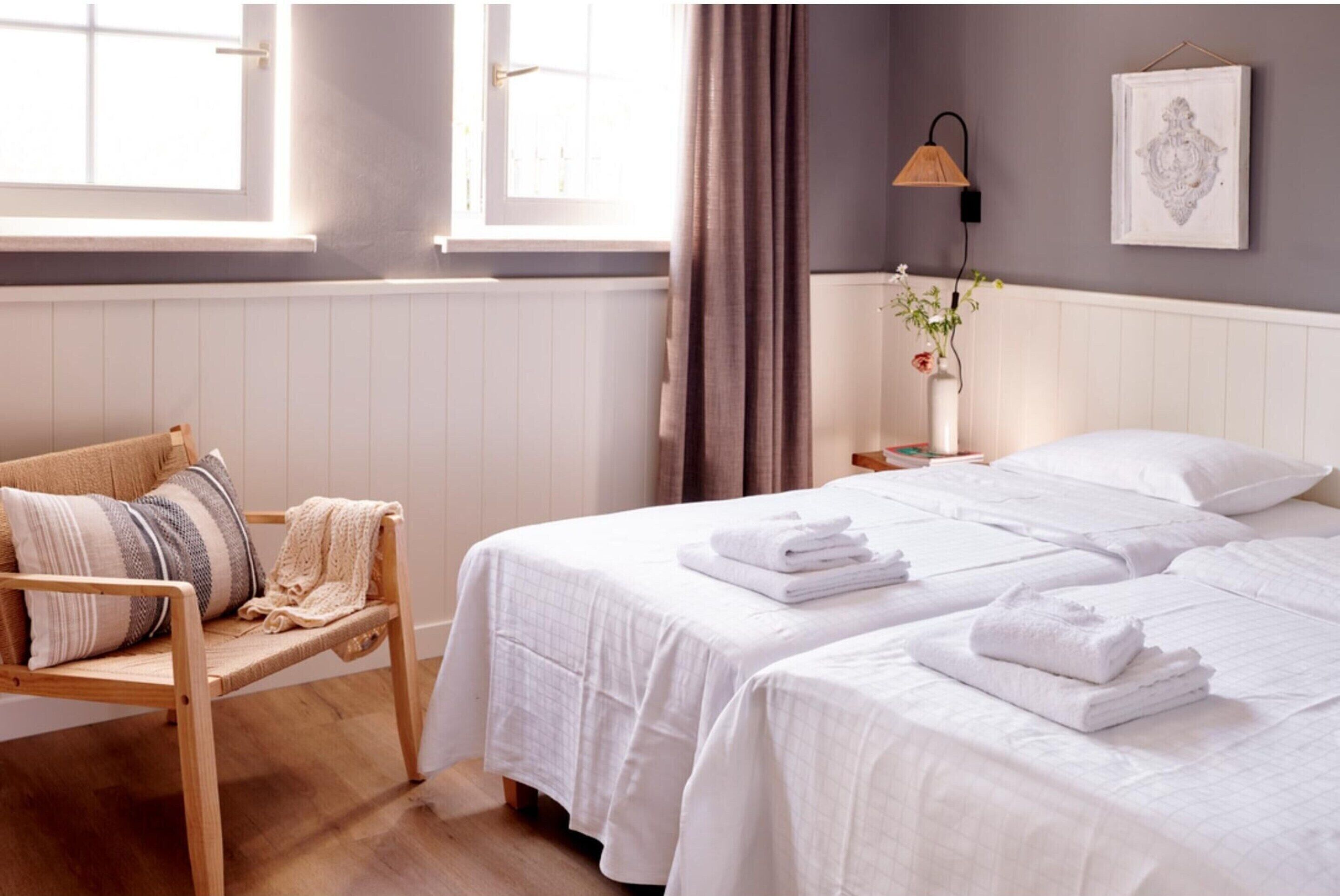 Een hotelkamer met twee bedden, een houten stoel, een nachtkastje met een lamp, een radiator en een raam met gordijnen.