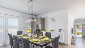 A modern dining area with a dark wooden table set for four, surrounded by grey upholstered chairs. A pendant light fixture hangs above the table, and a kitchen area is visible in the background. - Villa in Novigrad With Private Pool (Novigrad)