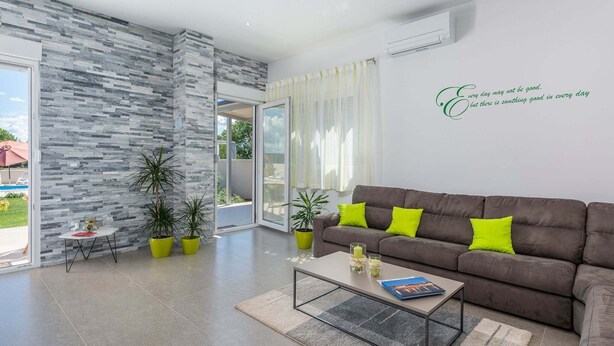 A modern living room with a grey stone wall, a brown sofa with bright green cushions, a glass coffee table, and a view of a pool through a glass door. - Villa in Novigrad With Private Pool (Novigrad)