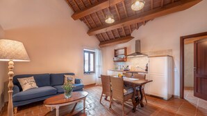 A cozy living room with a blue sofa, a wooden coffee table, and a dining table with wicker chairs. A kitchen is visible in the background with a window and a white refrigerator. - Serene Apartment With Pool (Mombaroccio)