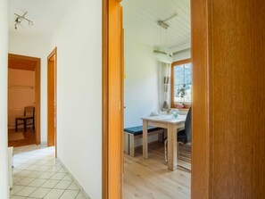 A room with a wooden floor, a table with a chair, a window with a view, and a door leading to another room.