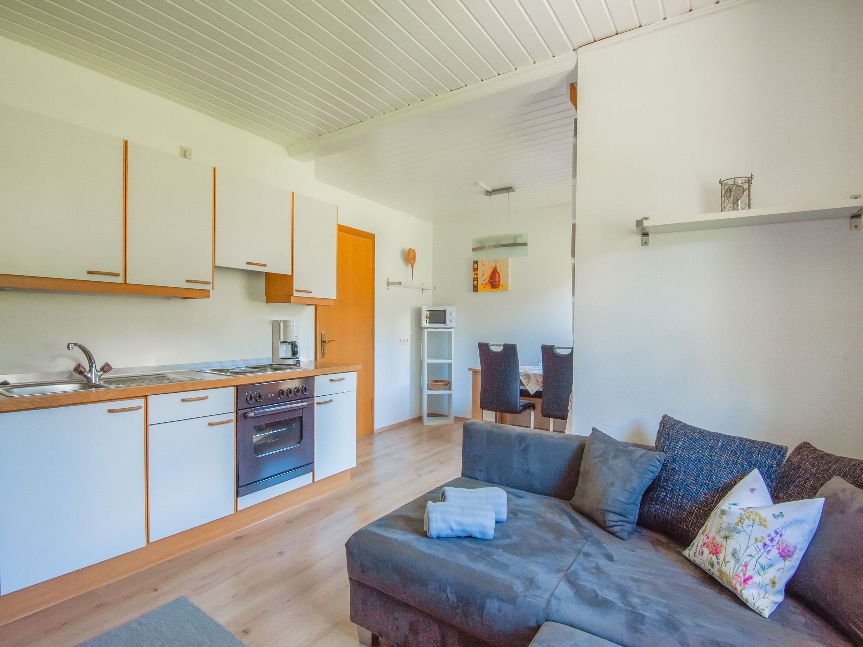 Una cocina moderna con armarios blancos, horno empotrado y microondas. Hay un comedor con mesa y sillas, y una sala de estar con sofá gris y cojines decorativos.