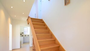 Un escalier en bois mène à l’étage supérieur, et on aperçoit une cuisine au fond.