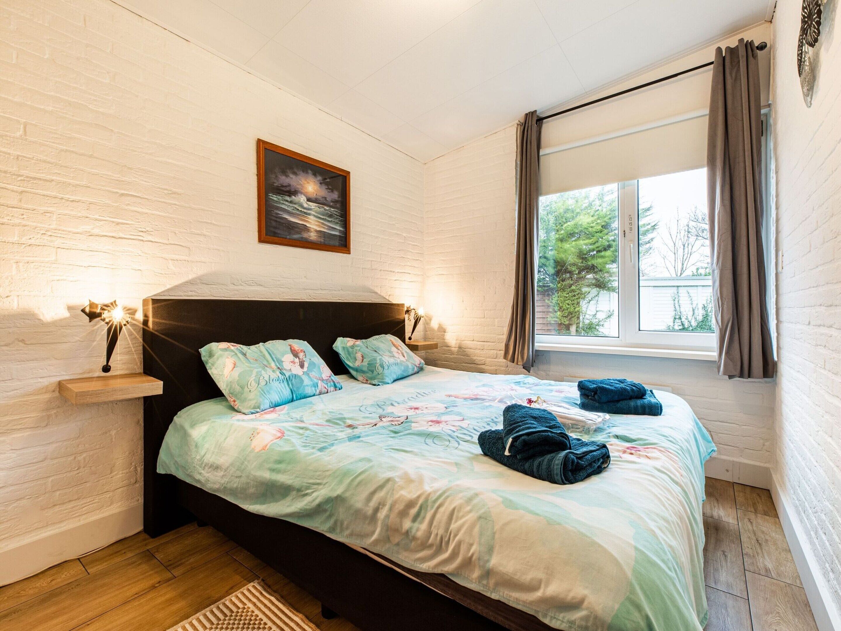 Een slaapkamer met een bed, een nachtkastje, een wandlamp, een raam met gordijnen en een ingelijst schilderij aan de muur.