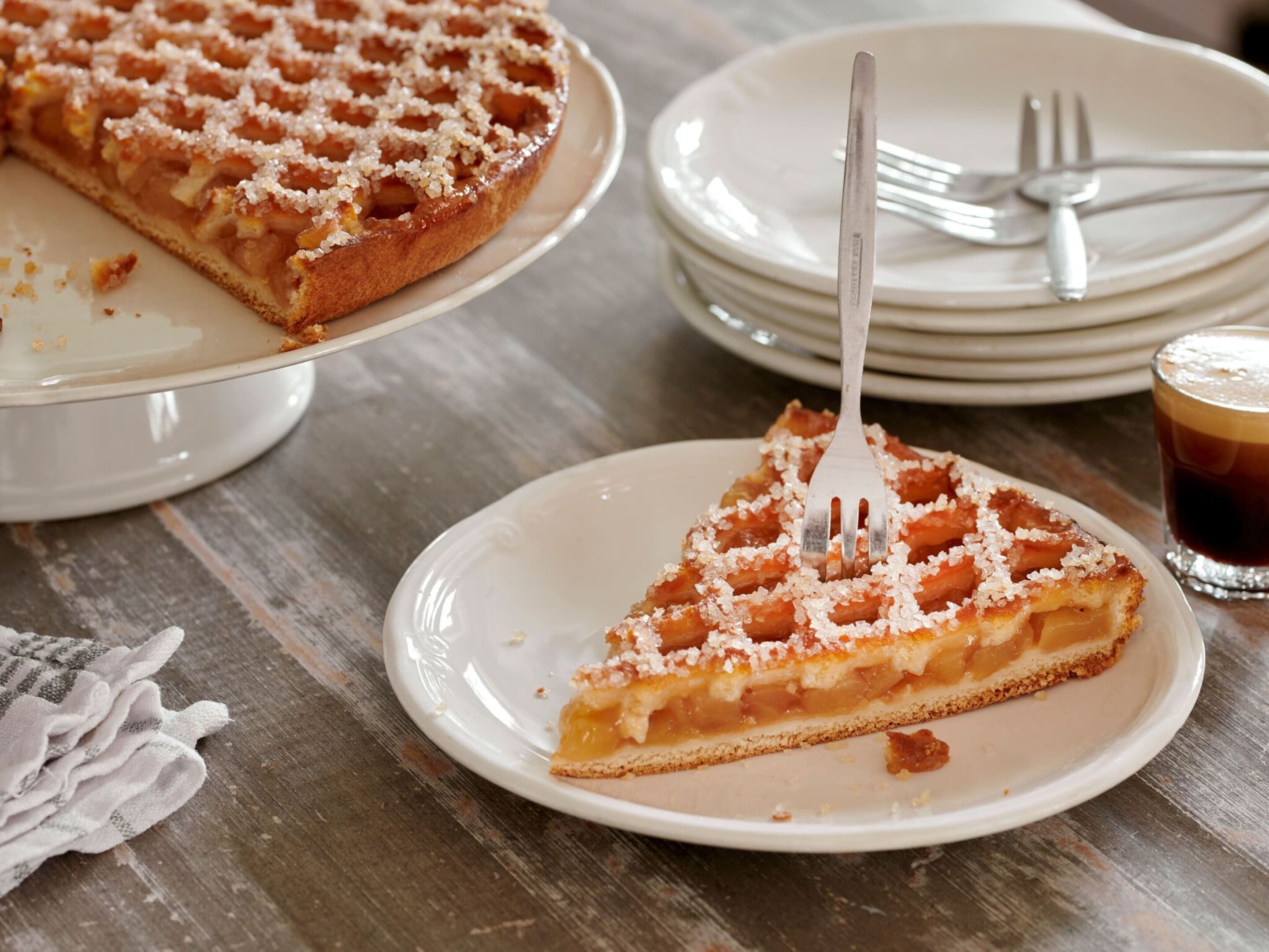 Una porción de tarta de manzana en un plato blanco con un tenedor, una tarta entera sobre un soporte, y una pila de platos con un vaso de café helado.