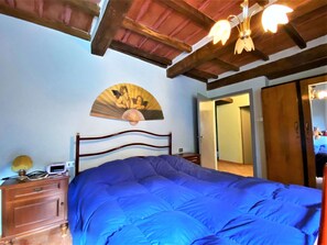 Ein Schlafzimmer mit einem großen Bett, einem Holz-Kopfteil, einem Nachttisch mit Lampe, einem Wandventilator und einer Tür zu einem weiteren Raum.