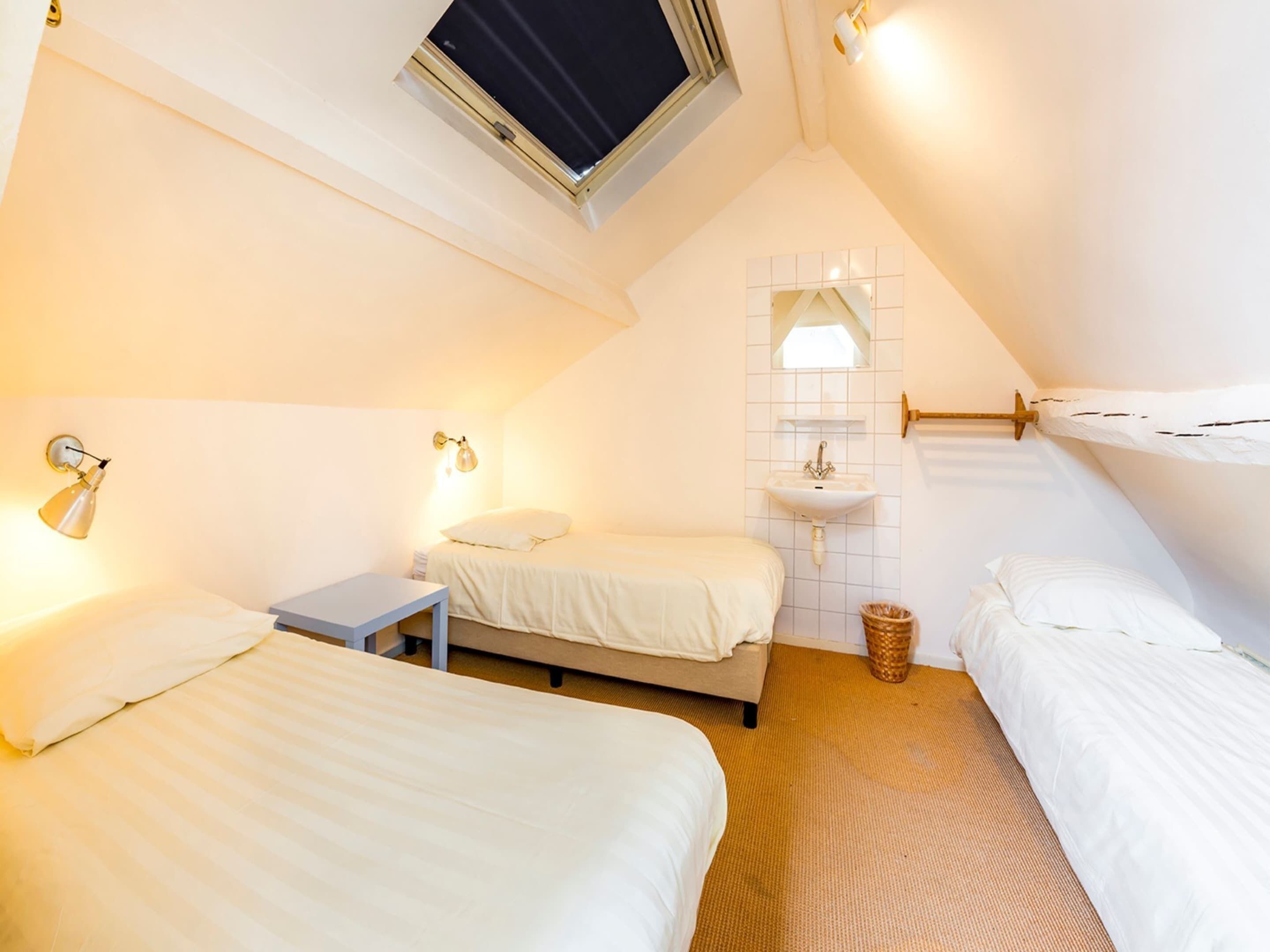 A compact hotel room with two single beds, a small bedside table, a wall-mounted sink, and a skylight.