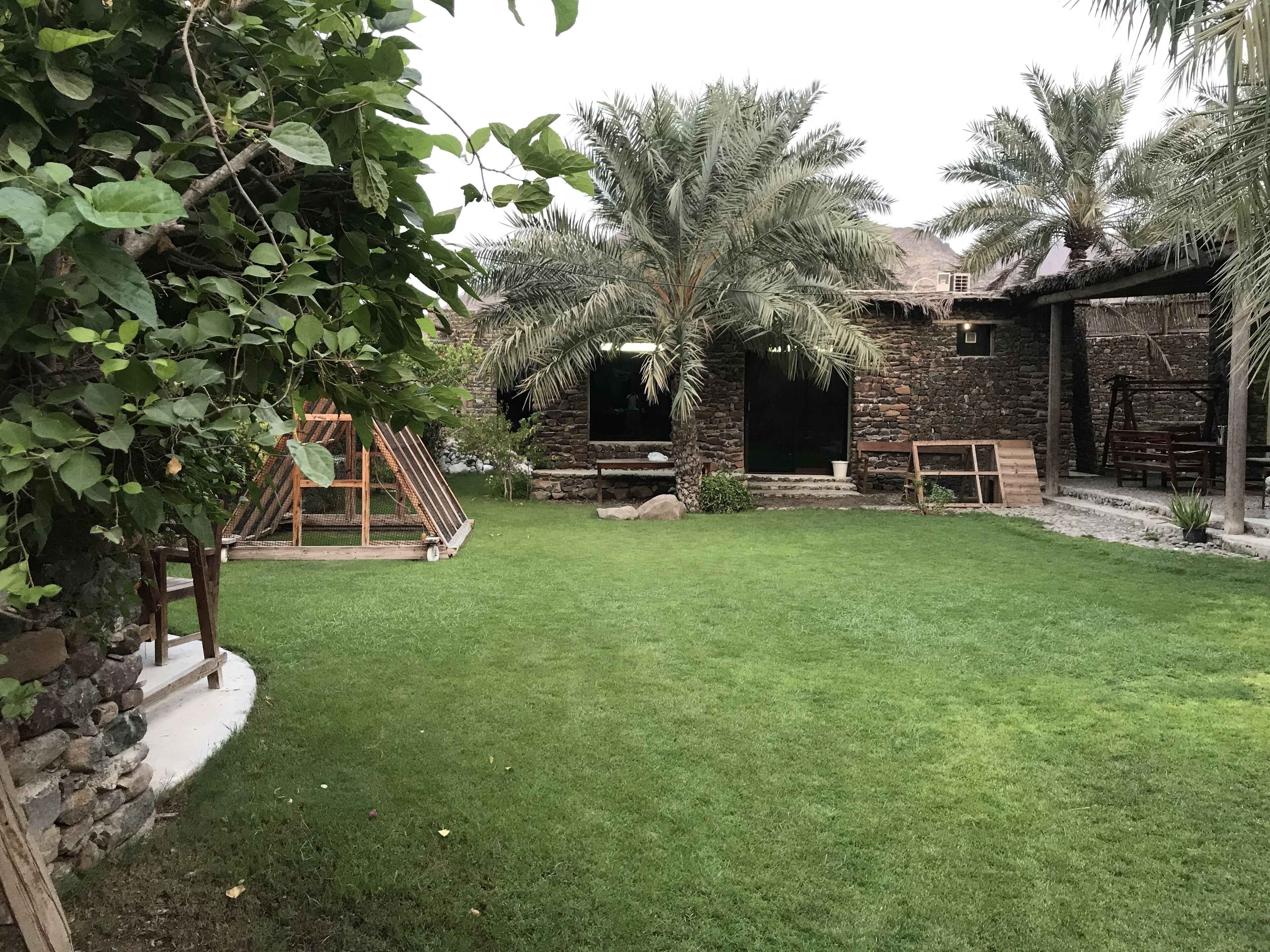 Un jardin bien entretenu, avec une pelouse verdoyante, une balançoire en bois et un bâtiment en pierre surmonté d’un toit de chaume.