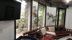 A cozy room with a wooden sofa, a small chair, and a TV mounted on the wall. - Among the Mountains (Mirbah)