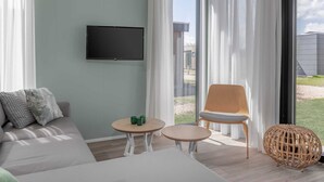 Un salon moderne avec un canapé, un téléviseur fixé au mur, une petite table ronde, un pouf en osier et une vue sur l’extérieur grâce à de grandes fenêtres.