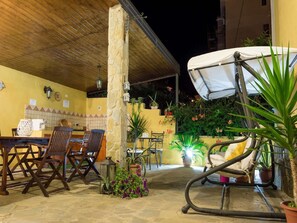 An outdoor patio area with a wooden table and chairs, surrounded by potted plants and a swing chair.