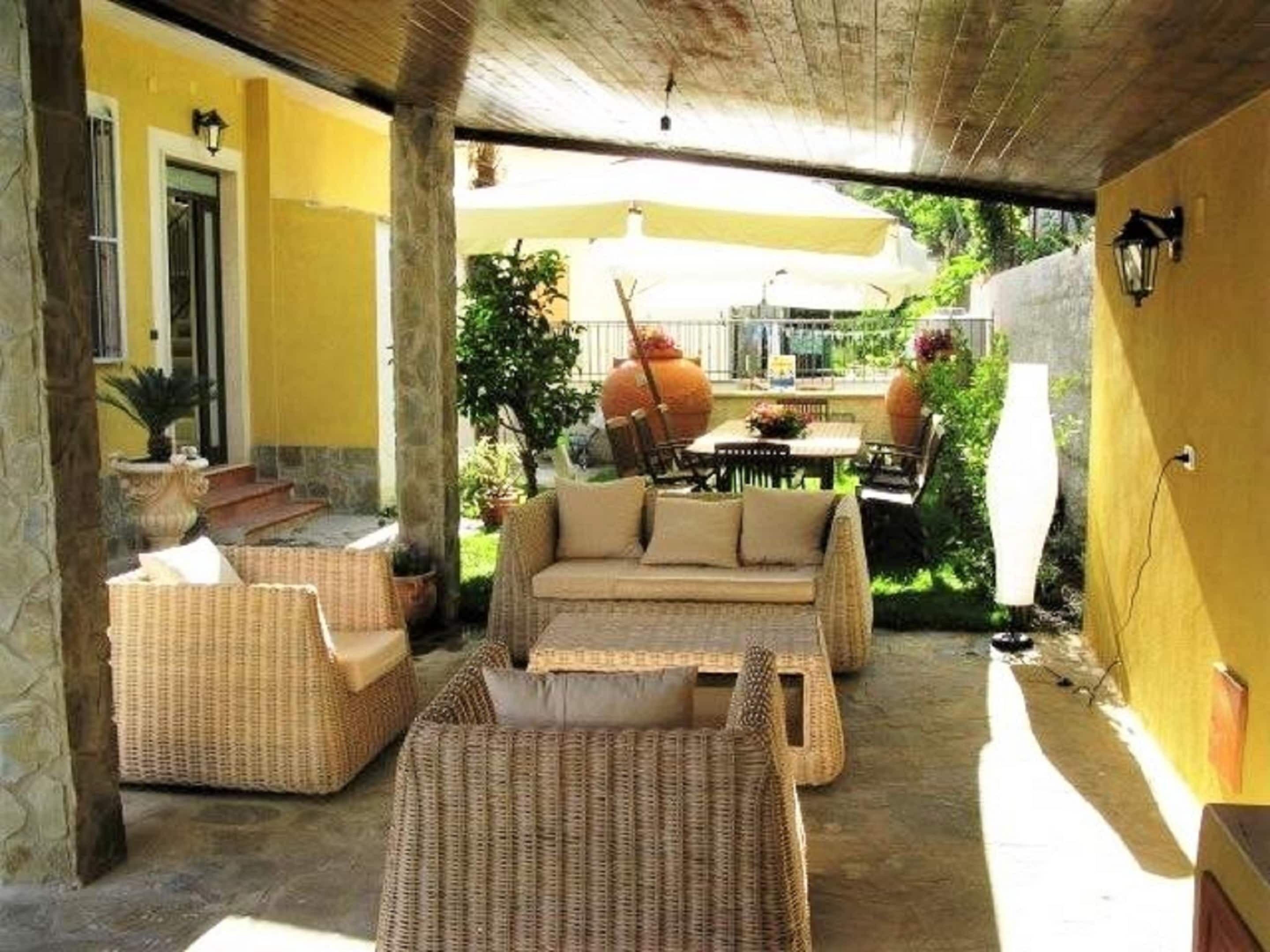 A covered outdoor seating area with wicker furniture, a table, and potted plants.