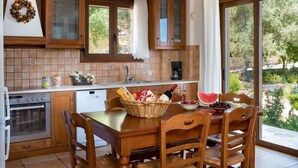 A kitchen with wooden cabinets, a tiled backsplash, a dining table with chairs, and a view of a garden through large windows. - Villa in Tavronitis With Pool & Stunning Views (Platanias)