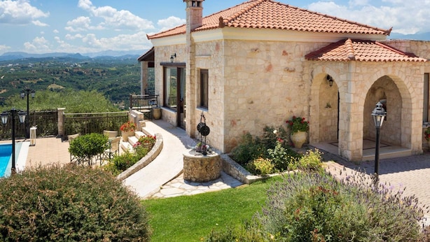 A stone building with a tiled roof, a swimming pool, and a garden with various plants and flowers. - Villa in Tavronitis With Pool & Stunning Views (Platanias)
