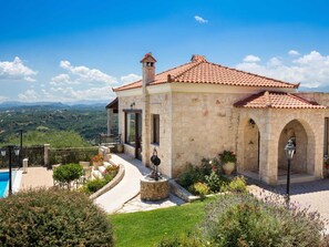 A stone building with a tiled roof, a swimming pool, and a garden with various plants and flowers.