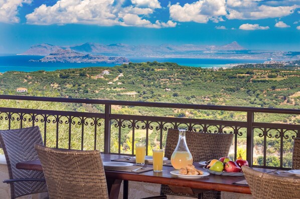 A balcony with wicker furniture, a table with plates, glasses, and a bottle, overlooking a scenic landscape with mountains and the sea.