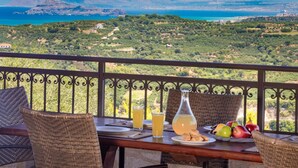 A balcony with wicker furniture, a table set with plates, glasses, and a pitcher of juice, overlooking a coastal landscape with mountains and greenery. - Villa in Tavronitis With Pool & Stunning Views (Platanias)