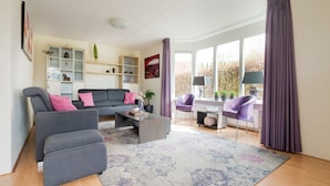 A modern living room with a grey sofa, a coffee table, and a patterned rug. There are purple chairs, a bookshelf, and a window with curtains. - Vacation Home With Jacuzzi (Schoonloo)