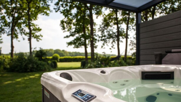 A hot tub with a control panel, surrounded by a wooden deck and a glass enclosure, with a view of a grassy area and trees in the background. - Vacation Home With Jacuzzi (Schoonloo)