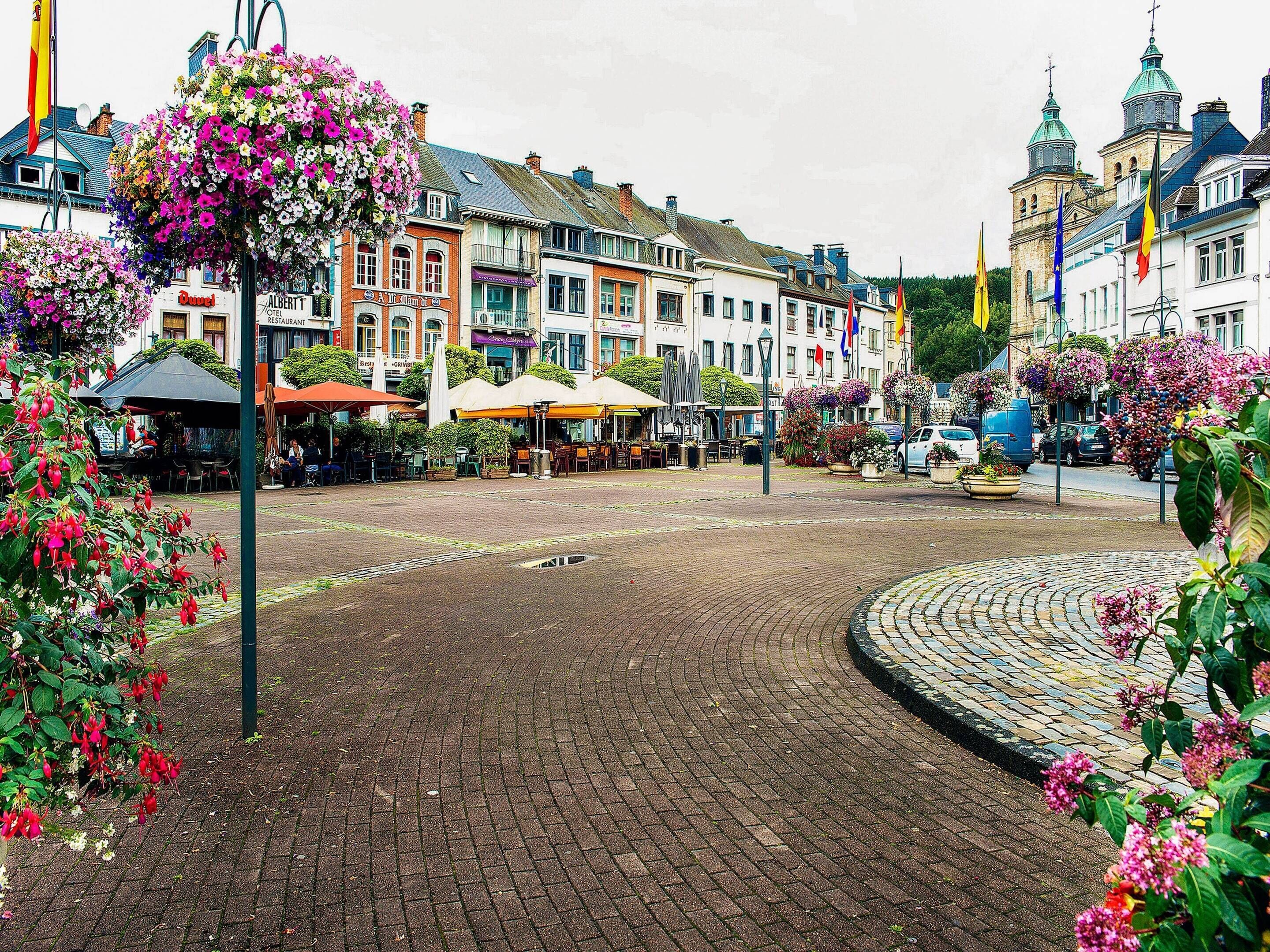 A cobblestone square with flower baskets, outdoor seating, and historic buildings with flags.
