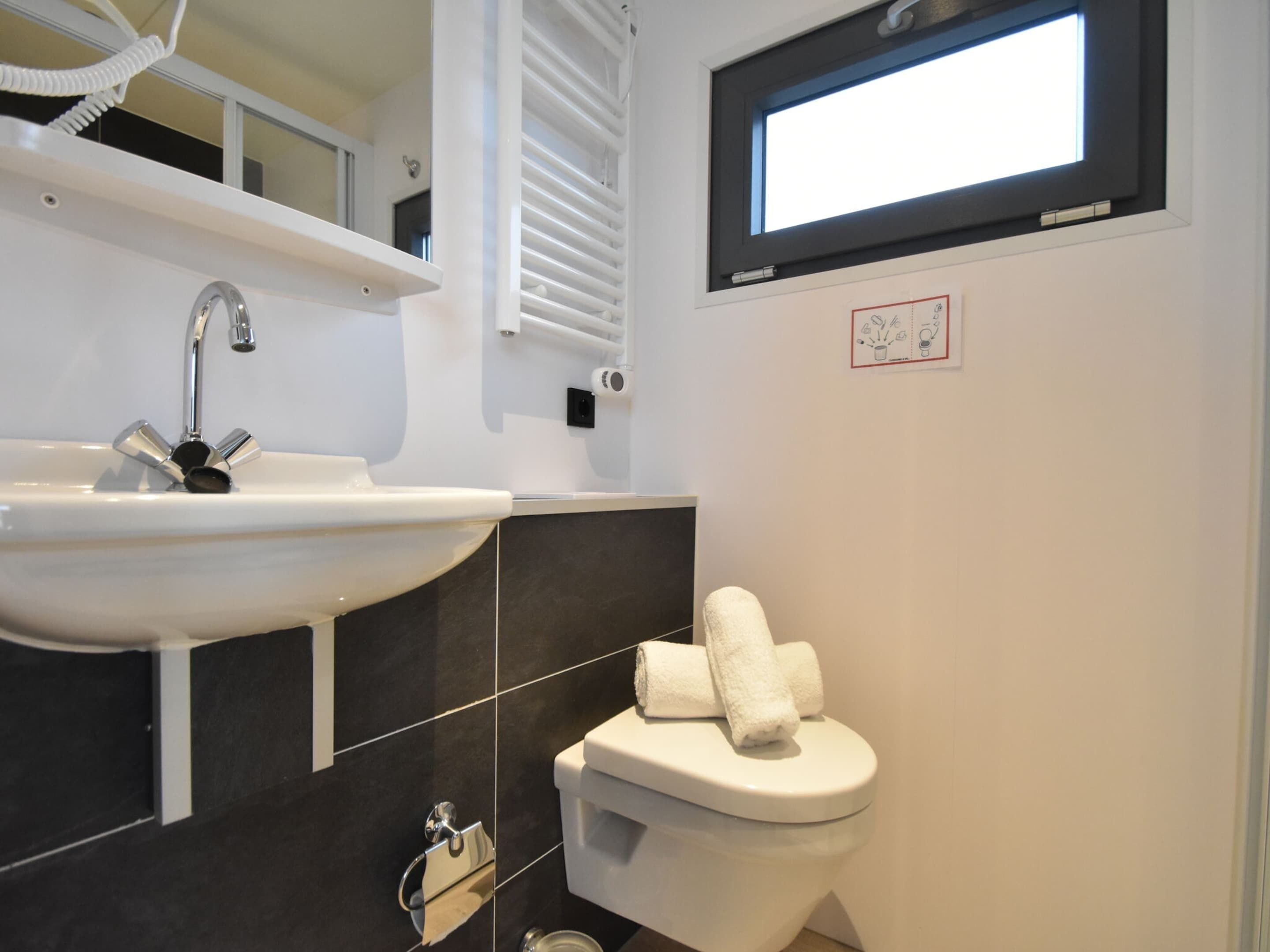 Ein modernes Badezimmer mit einem weißen Waschbecken, schwarzen Fliesen, einer Toilette und einem Fenster über der Toilette.