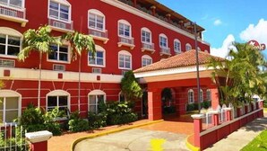 A red building with multiple windows and balconies, a tiled roof, and a paved entrance area with a yellow curb. - Hotel Seminole (Managua)