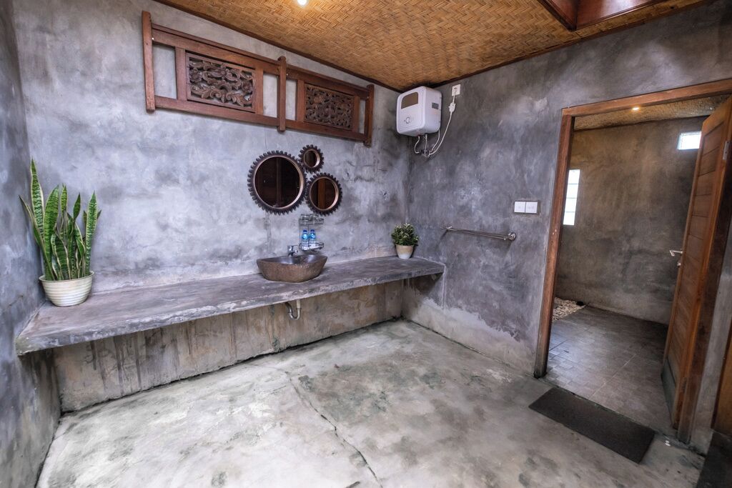 Baño con piso de concreto, repisa de madera con lavabo y placas decorativas en la pared, además de una planta en maceta.