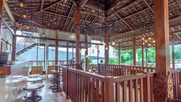 Un vaste espace intérieur doté de poutres en bois et d’un haut plafond, comprenant de nombreuses tables et chaises, de grandes fenêtres et une vue sur un paysage naturel.