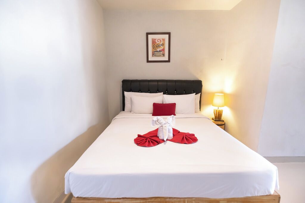 Una cama bien hecha con sábanas blancas, un almohadón rojo y una toalla blanca dispuestas sobre la cama. Hay un cuadro enmarcado en la pared sobre la cama y una lámpara de noche a la derecha.