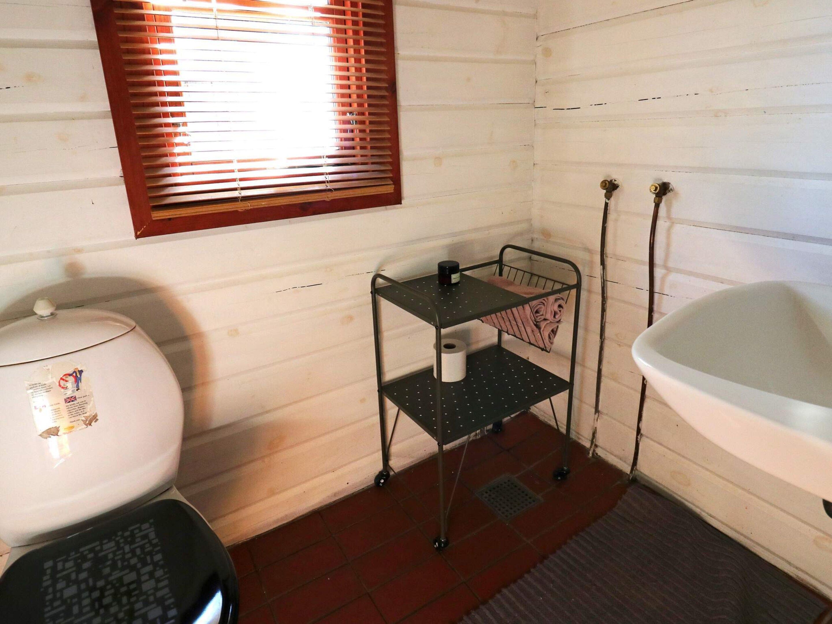 A compact bathroom with a toilet, a small metal shelf with a roll of toilet paper, a window with blinds, and a bathtub.