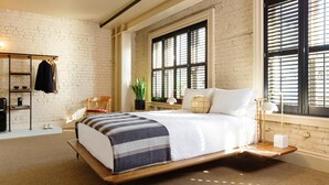 A modern bedroom with a bed, a wooden bench, a shelf with clothing, and a window with blinds.
