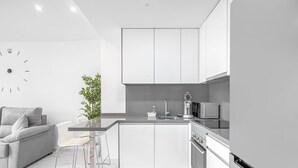 Une cuisine moderne avec des meubles blancs, un sol en marbre, un coin repas avec une table et des chaises en verre, et un canapé gris.