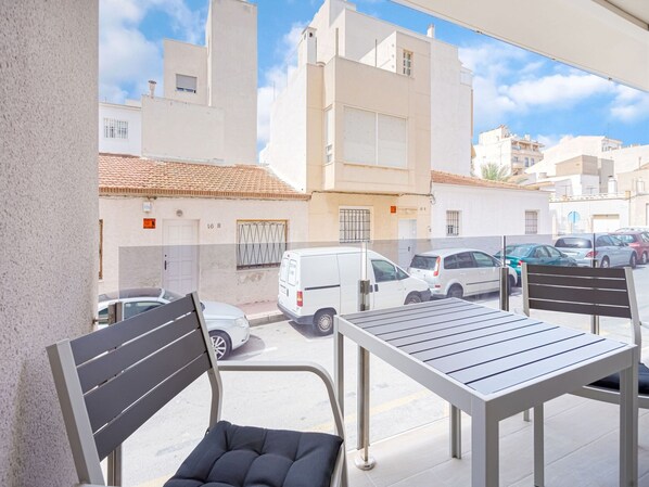 Un balcón con mesa y sillas, con vista a una calle con coches aparcados y edificios.