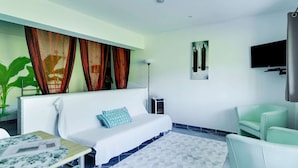 Ein modernes Hotelzimmer mit einem weißen Sofa, einem grünen Sessel, einem kleinen Tisch mit einem Dekorationsobjekt und einem großen Fenster mit roten Vorhängen.