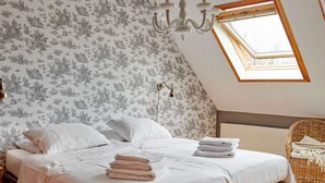 Een slaapkamer met een bed, kussens, een stapel handdoeken, een kroonluchter, een raam en een rieten stoel.